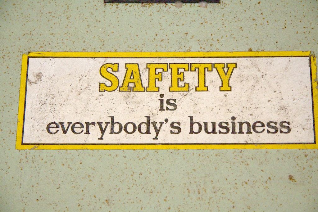 Photograph of a safety sticker on a green wall. The sticker is rectangular and white with a yellow border. The word ‘Safety’ is written in yellow capitalised text, with ‘is everybody’s business’ written underneath in lower case black.