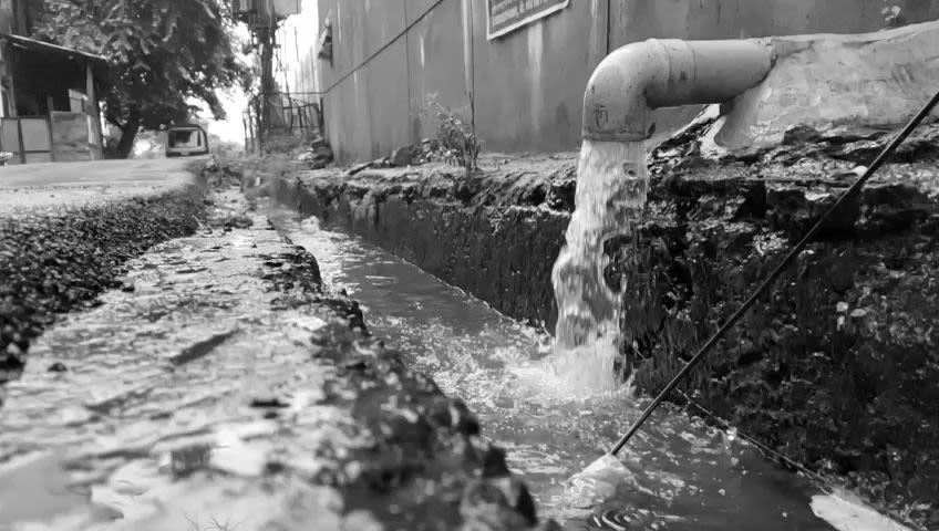 Black and white photograph of a ground-level water pipe spilling into a drain beside a road. There is a building visible on either side of the road at the top of the picture.  