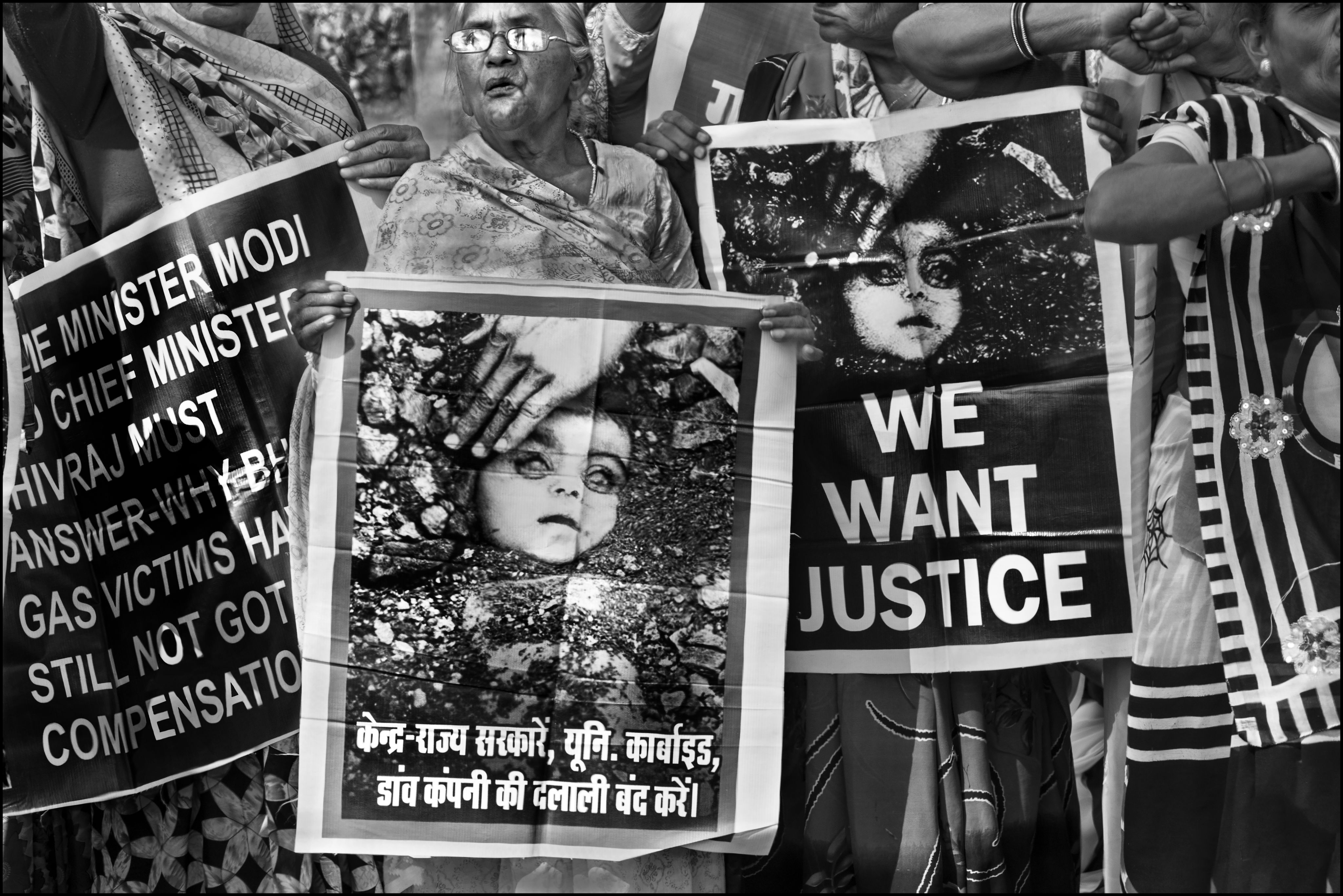 Activists holding posters at a demonstration; their faces are not visible. The posters include Rai's photograph 'Burial of an Unknown Child' with text in Hindi and English. The English text reads 'We want justice'.