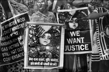 Activists holding posters at a demonstration; their faces are not visible. The posters include Rai's photograph 'Burial of an Unknown Child' with text in Hindi and English. The English text reads 'We want justice'.