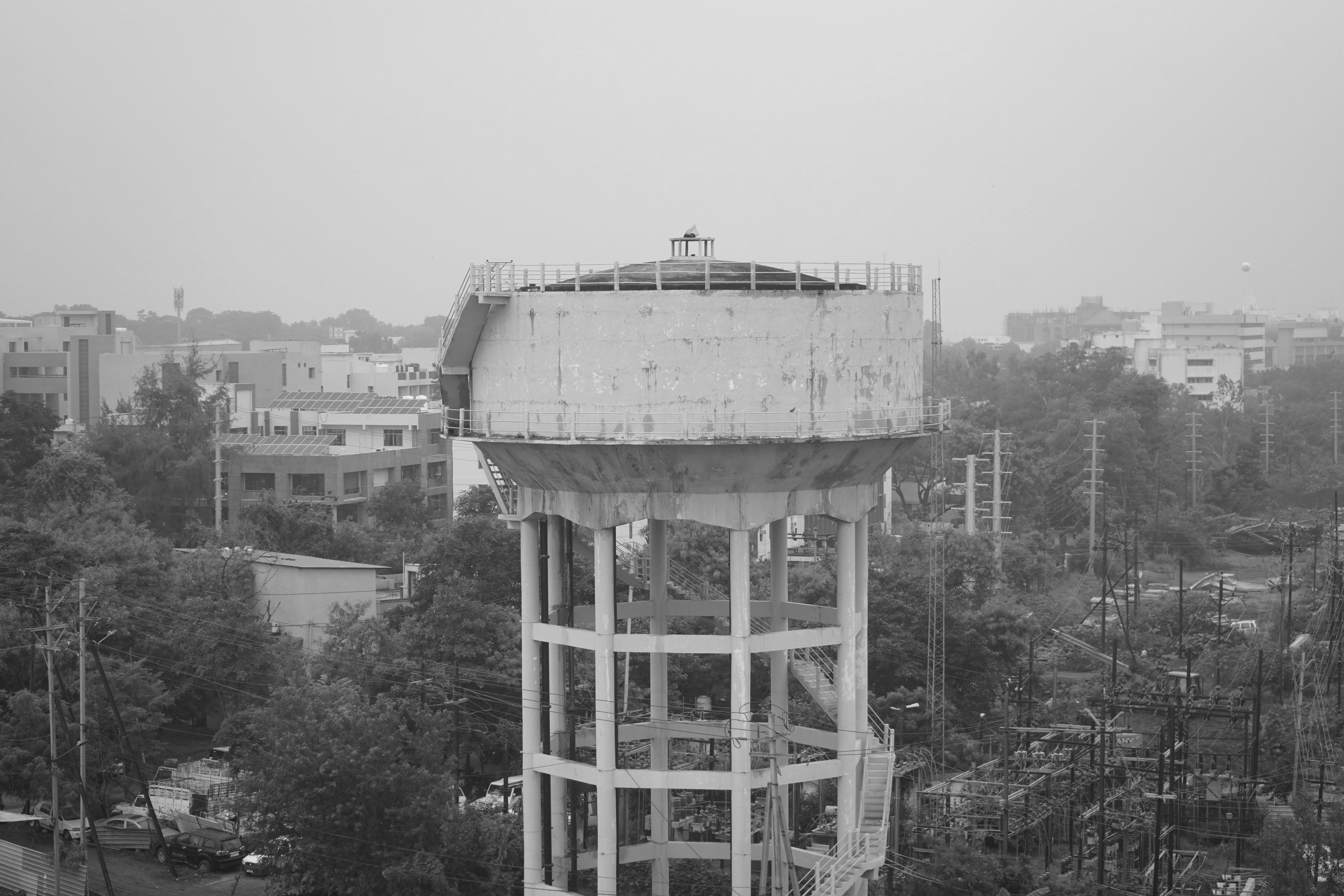 Black and white photograph of a very large round concrete tank on a high stilt-like structure, rising above trees and buildings. There are steps up to the tank and a fenced off platform for walking around its base and top.